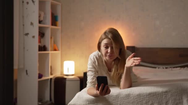 パジャマの若い金髪の女性は 愚かな対話者によって複雑にスマートフォン携帯電話を話すベッドの上に横たわる — ストック動画