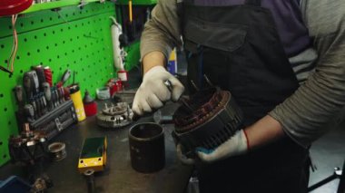 Tamirci elinde bir araba jeneratörü ya da elektrikli bir motor tutuyor ve cırcırlı alet kullanarak cıvataları ve fındıkları özenle çeviriyor.