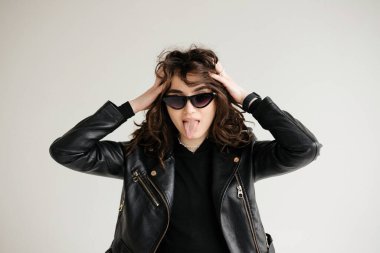 Brunette with curly hair, glasses and leather jacket shows her tongue isolated. High quality photo