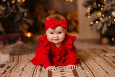Kırmızı elbiseli ve bantlı mutlu bir bebek Noel ağacının önünde oturuyor. Gülümsüyor ve doğayla iç içe eğleniyor. Sevimli bebek kıyafetleri giyerken.