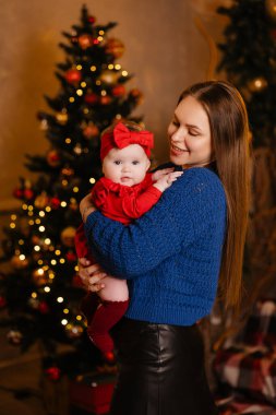 Bir kadın Noel ağacının önünde duruyor, yüzünde bir gülümseme olan bir bebeği tutuyor. Bebek kafasına şirin bir Noel süsü takmış ve ikisi de mutlu ve neşe dolu görünüyor.