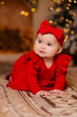 Kırmızı elbiseli ve bantlı mutlu bir bebek Noel ağacının önünde oturuyor. Gülümsüyor ve doğayla iç içe eğleniyor. Sevimli bebek kıyafetleri giyerken.