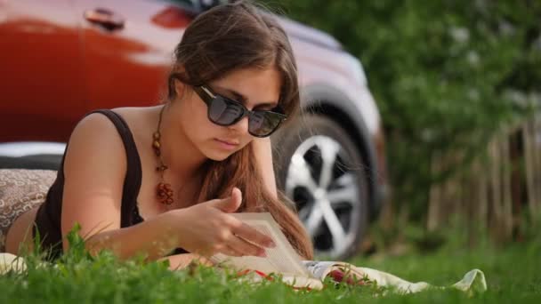 一个戴着太阳镜的年轻的黑发女人在车尾的草坪上看书 — 图库视频影像