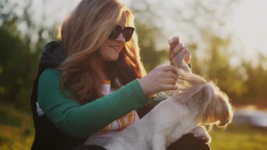 Güneş gözlüklü genç bir kadın parkta çimlerin üzerinde oturuyor, kollarında bir Shih Tzu köpeği tutuyor. Çevrelerinde yaprak döken ağaçlar ve gün batımı vardır. Parlak ve ılık bir atmosfer yaratırlar..