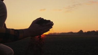 Bir adamın ellerine yaklaşan tarım uzmanı gün batımında bir tarlada elindeki toprağı inceliyor. Doğal organik toprak tarımı