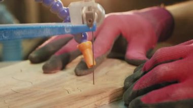 Eldivenli bir el, bant testeresi üzerinde çalışırken tahta bir tahta tutuyor. Tahtadan karmaşık bir desen kesme işlemi görülebilir.