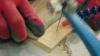 Eldivenli bir el, bant testeresi üzerinde çalışırken tahta bir tahta tutuyor. Tahtadan karmaşık bir desen kesme işlemi görülebilir.