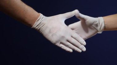 Beyaz lateks eldivenli iki el lateks bir tokalaşmada koyu mavi arka plana yapıştırılır. Bu, koruma gerektiren durumlarda güvenlik, hijyen ve işbirliğini sembolize eder.