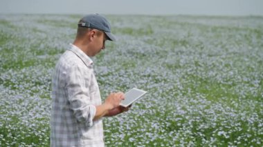 Damalı gömlekli ve şapkalı bir adam elinde tabletle açan beyaz ve mor çiçeklerle dolu bir tarlada duruyor. Ekrana odaklanmış, dışarıda çalışıyor.