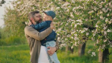 Başarılı, mutlu bir adamın portresi. Kollarında küçük oğlu çiçek açmış bir elma bahçesinde kameraya bakıp gülümsüyor.