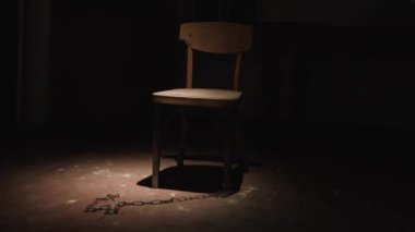 Odanın ortasında, dar bir ışık demeti ile aydınlatılmış, eski bir ahşap sandalye. Sandalyelerden birine zincir bağlanır. Bacakları izolasyon ve hapsetme ortamı yaratır.