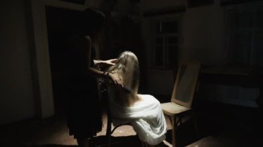 Karanlık bir odada iki genç kadın. Bir tanesi, uzun saçlı, beyaz bir beze sarılı bir sandalyede oturuyor. Diğeri yakınlarda duruyor, oturan kadınların saçlarını tarıyor.
