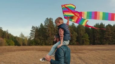 Bir adam küçük bir çocuğu omuzlarında taşıyor ve çocuk renkli bir uçurtma tutuyor. Açık bir alanda, açık mavi gökyüzünün altında bir ormanın yanındalar.