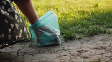 Bir insan yürürken mavi plastik torba kullanarak köpek atıkları topluyor. Süreç, yeşil çimenlerle çatlamış zeminde gerçekleşiyor.