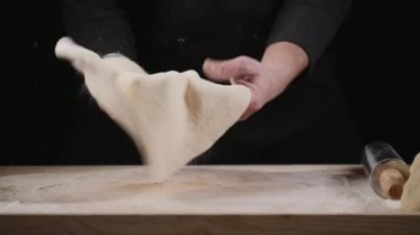 Tahta bir masanın üzerine hamuru fırlatan bir adamın elleri. Yanında da metal bir raptiye vardı. Hamur hareket halinde, arka plan karanlık.