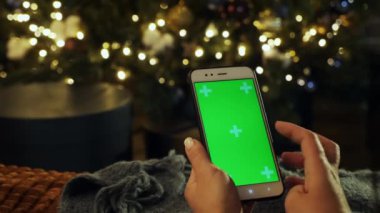 Akşam evdeki telefon ekranında kadın elinin yeşil ekrana dokunuşunun yakın çekimi. Krom anahtar, takip hareketi. Arka planda Noel ışıkları yanar.