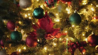 Parlak, çok renkli mücevherlerle, kırmızı çiçeklerle ve küçük ışıklarla süslenmiş bir Noel ağacı. Işıklar, ağaçtaki süslerden yansıyan ılık ve şenlikli bir hava yaratıyor.