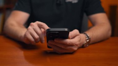 Siyah gömlekli bir adam ahşap bir masada oturuyor, elinde bir akıllı telefon tutuyor. Cihazı aktif olarak kullanıyor, ekrana odaklanıyor.