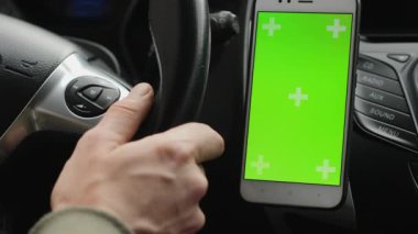 Bir el, yanında bir tutacağa monte edilmiş krom renkli yeşil ekran içeren akıllı telefonlu bir araba direksiyon simidini tutuyor. Arabanın kontrol panelinin bir kısmı görünüyor.