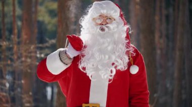 Neşeli bir Noel Baba 'nın yakın plan portresi kameraya bakar ve Noel arifesinde güneşli kış havasında parkta dikilirken parmağını kaldırır.