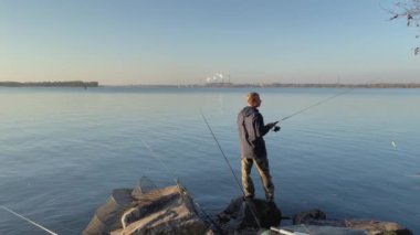 Bir balıkçı, gölün kıyısında sigara tüten endüstriyel borular ve gölet kirliliği ile çalışan bir fabrikanın arka planına karşı durarak balık yakalar.