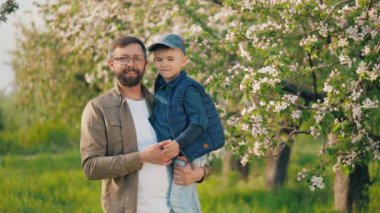 Başarılı, mutlu bir adamın portresi. Kollarında küçük oğlu çiçek açmış bir elma bahçesinde kameraya bakıp gülümsüyor.