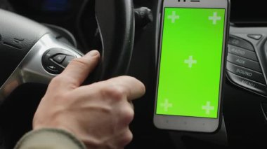 Bir el, yanında bir tutacağa monte edilmiş krom renkli yeşil ekran içeren akıllı telefonlu bir araba direksiyon simidini tutuyor. Arabanın kontrol panelinin bir kısmı görünüyor.