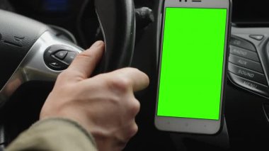 Yakından çekilen sürücü direksiyon tutarken araba sürüyor, gösterge panelinde yeşil ekranlı bir akıllı telefon var. Mobil uygulama teknolojisi ve navigasyon