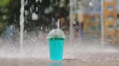 Mavi kokteyli olan plastik bir bardak çeşmenin önünde ıslak bir yüzey üzerinde duruyor. Arkaplan, su damlacıklarının etrafa saçıldığı bulanık ağaç ve bina hatlarına sahip.