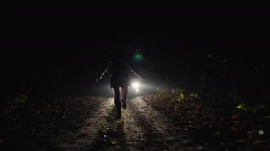 Karanlık bir yolda arabanın farlarına doğru koşan bir insanın silüeti. Işıklandırma, gece sahnesi için dramatik bir atmosfer yaratır.