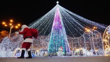 Sırtında kırmızı bir torbayla Noel Baba gece vakti parlak bir Noel ağacının önünde yürür.