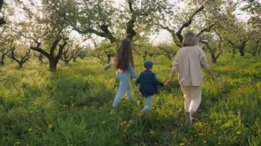 Baharda, çiçek açan ağaçlar ve canlı çimlerle çevrili yeşil bir elma bahçesinde el ele tutuşan bir aile yürüyor. Güneş ışığı dalların arasından süzülür