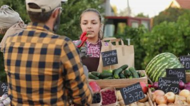 Çiftçi pazarında bir kız müşteriye kırmızı biber gösteriyor, etrafında sebzeler var.