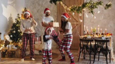 Noel şapkalı ve ekose pijamalı bir aile tatil süslemeleri, Noel ağacı ve mumlarla dolu bir şenlik masasında birlikte vakit geçiriyorlar.