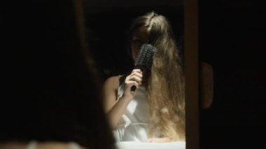 Bir kız karanlık bir odada aynanın önünde uzun saçlarını tarıyor, yumuşak ışıkla aydınlatılıyor, sakin ve içsel bir atmosfer yaratıyor.