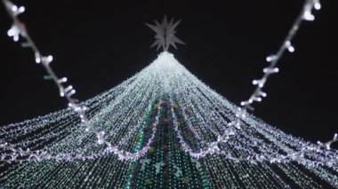 Parlayan çelenklerle süslenmiş bir Noel ağacının tepesi ve gece gökyüzüne bakan bir yıldız.