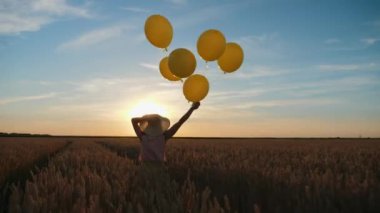 Buğday tarlasında gün batımına karşı sarı balonlar tutan bir kız.