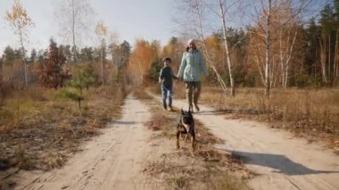 Sonbahar ormanlarında kumlu bir yolda oğluyla ve köpeğiyle yürüyen bir anne. Altın yapraklı ağaçlarla çevrili güneşli bir gün.