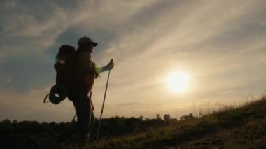 Sırt çantalı bir kadın yürüyüşçü silueti ve gün batımında yürüyüş direkleri, yokuş yukarı yürüme