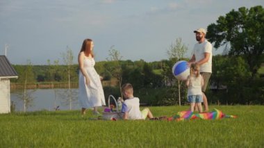 Aile, yaz günü göl kenarındaki yeşil bir çayırda vakit geçiriyor. Baba kızıyla top oynar, anne izler ve oğul piknik sepetinin yanına oturur.