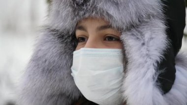Yumuşak başlıklı ve koruyucu tıbbi maskeli bir kız kışın dışarıda duruyor. Bakışları sakin ve arka planı karlı.