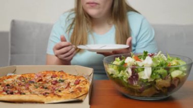 Önünde taze yeşil bir salata kasesi ve pizza kutusuyla bir kadın yemek için hazırlanıyor. Atmosfer rahat, sade ve rahat.