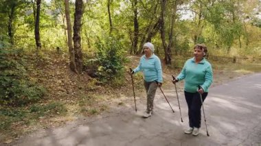 İki yaşlı kadın bir orman yolunda İskandinav kutuplarıyla yürüyor. Aktif yaşam tarzı, açık hava fitness ve yaşlılar için sağlık