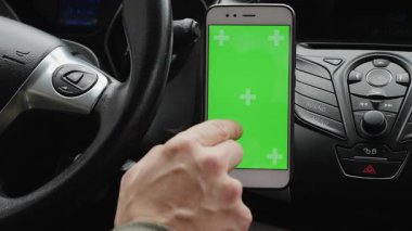 Arabada otururken elinde yeşil ekran olan akıllı telefonu tutan birinin yakın çekim görüntüsü. Telefon, gösterge paneli ve direksiyonun önüne, ekranı gösteren bir parmak ile yerleştirilmiş.