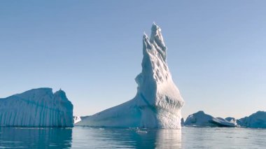 Sakin suların üzerinde güneş ışığını yansıtan devasa bir buzdağı kuleleri. Serin renkler bu açık günde kutup ortamının güzelliğini vurgular..