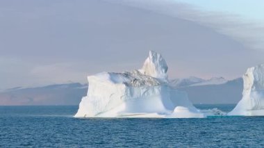 Büyük bir buzdağı, sabahın erken saatlerinde berrak mavi sularda kayarak uzak dağlara karşı eşsiz şeklini gösterir..