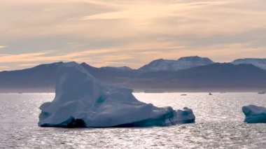 Büyük bir buzdağı, sabahın erken saatlerinde berrak mavi sularda kayarak uzak dağlara karşı eşsiz şeklini gösterir..