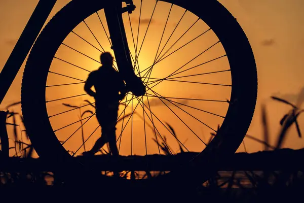 Close-up silhouette of a bike wheel at sunset. The sun shines through the wheel of a bicycle with blurred silhouette of running man, selective focus