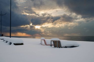 Karla kaplı plajda, bulutlu gün batımı arkaplanlı iki bank.