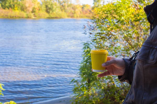 Man holding yellow cup on river background. Coffe cup in nand on autumn landscape background. Travel concept. Vacations at nature. Coffee to go concept. Picnic at the riverbank. Active morning.